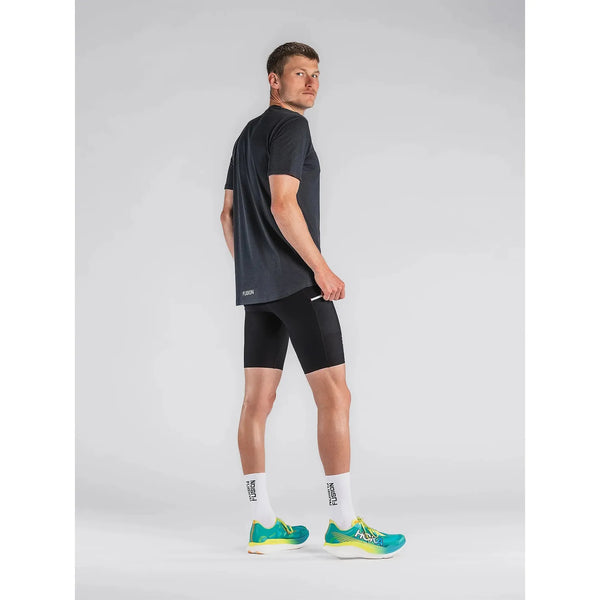 En mand står i et studie iført Fusion Short Tights Advanced, Unisex, Superblack, sammen med en sort T-shirt, hvide træningssokker med sort tekst og blågrønne løbesko med gule detaljer. Han kigger væk og ser sig over skulderen.