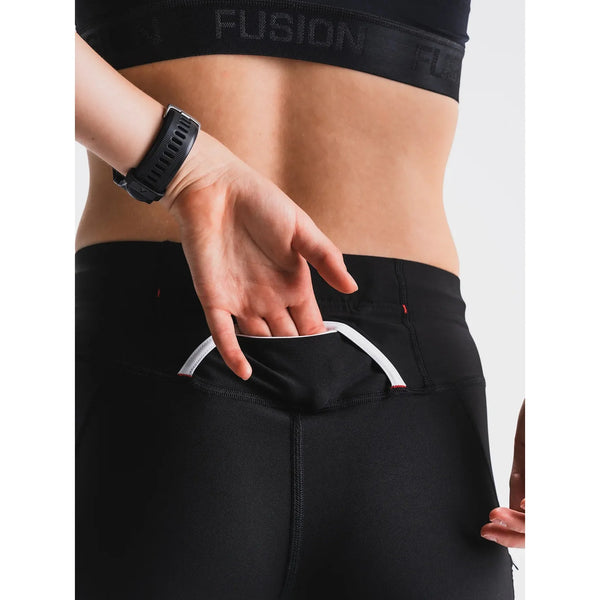 Iført Fusion's Short Tights Advanced, Unisex, Superblack, bruger en person sin højre hånd til at åbne en lille baglomme i linningen, som afslører en hvid foring - ideel til opbevaring af vigtige ting, mens man løber.