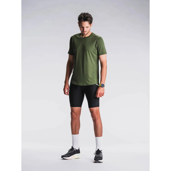 En mand står på en almindelig lys baggrund iført en grøn kortærmet skjorte, Fusion Short Tights Advanced Unisex i Superblack, hvide sokker og sorte løbesko. Han kigger ned med armene afslappet langs siden.