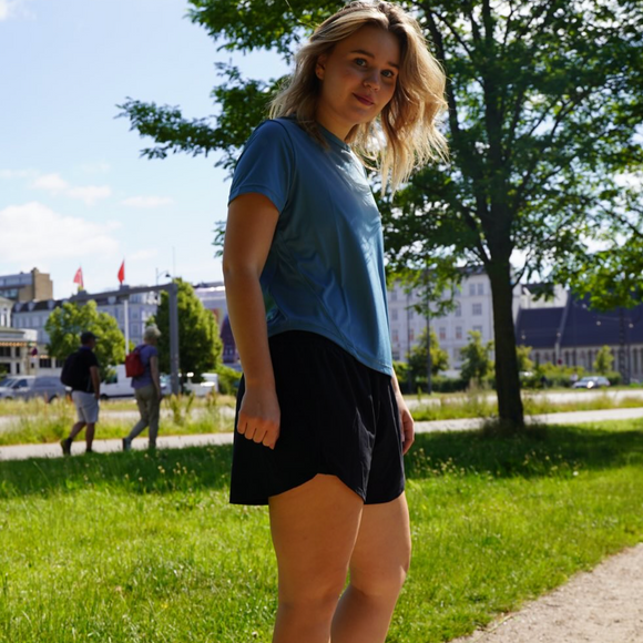 En person i blå skjorte og sorte shorts står på græs i en solrig park med træer, bygninger og to mennesker, der går i baggrunden.