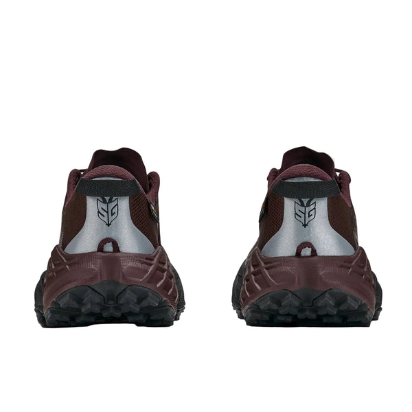 Et par Hoka Speedgoat 7 GTX trailsko til kvinder i farverne Deep Mauve og sort er afbilledet set bagfra på en hvid baggrund. Skoene har strukturerede såler og et reflekterende, sølvfarvet, geometrisk logo på hælen. De er fremstillet med GORE-TEX og yder fremragende beskyttelse i vådt vejr.