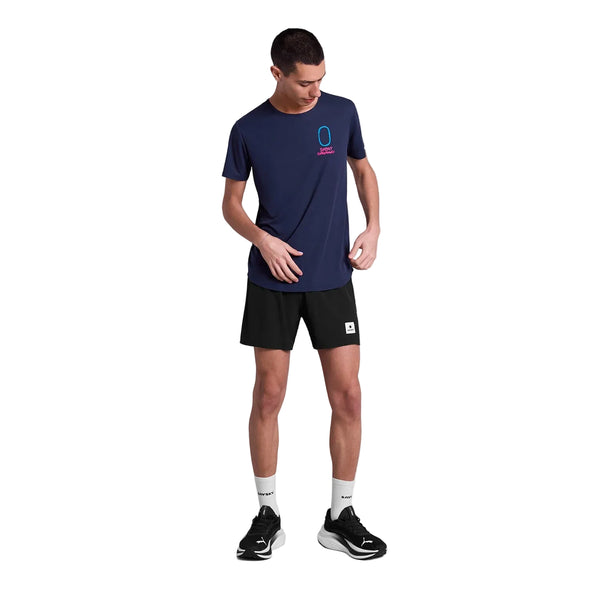 En mand i en marineblå Statement Flow T-shirt til mænd fra Saysky, sorte shorts, hvide sokker og sorte sportssko står på en hvid baggrund, mens han kigger ned og retter på sin skjorte.