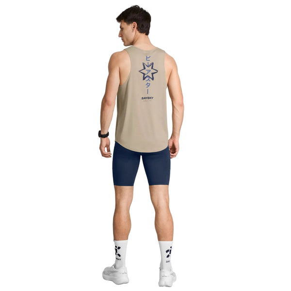 En mand står med ryggen til, iført en Saysky Statement Combat Pro Singlet, Herre, Sand – med et blåt motiv og tekst – kombineret med marineblå sportsshorts, hvide strømper med mørkeblå mønstre og hvide sneakers. Hans ærmeløse top er åndbar og hurtigtørrende, hvilket sikrer komfort under intense træningspas. Han har kort brunt hår og et ur på venstre håndled.