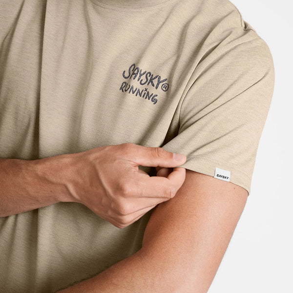 En person trækker i ærmet på en sandfarvet Statement Combat Pro T-shirt, Herre fra Saysky, med teksten »SAYSKY® RUNNING« trykt på brystet og et lille Saysky-mærke på ærmet. Kun personens overkrop og arm er synlige på en ensfarvet baggrund.