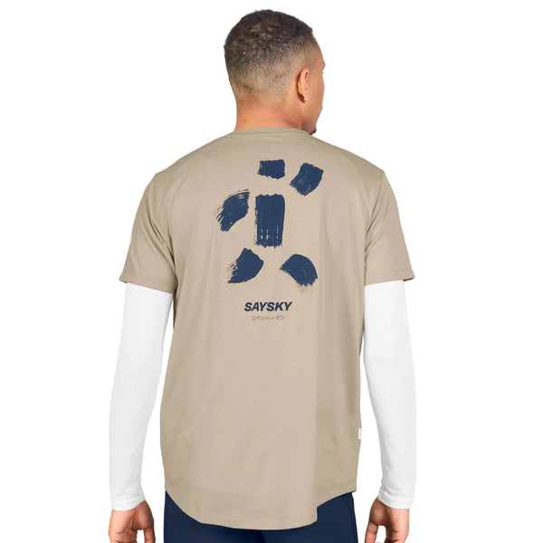 Der ses en person set bagfra, iført en Saysky Statement Combat Pro T-shirt, Herre, Sand – en beige, hurtigtørrende løbetrøje til mænd – over en hvid langærmet skjorte. T-shirten er prydet med abstrakte, marineblå penselstrøgsmønstre og teksten »SAYSKY« trykt på ryggen.