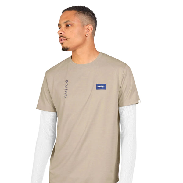 En mand iført Saysky Statement Combat Pro T-shirt, Herre, Sand – en beige kortærmet T-shirt med et lille marineblåt mærke – over en hvid langærmet trøje, står foran en ensfarvet hvid baggrund og kigger mod venstre. Denne hurtigtørrende løbetrøje til mænd fra Saysky kombinerer både komfort og stil til enhver aktiv dag.