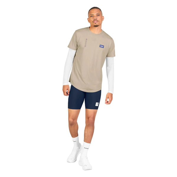 En mand står foran en hvid baggrund iført en Saysky Statement Combat Pro T-shirt, Herre, Sand, som er trukket uden på en hvid langærmet skjorte, kombineret med marineblå shorts, hvide ankelstrømper og hvide sneakers. Denne hurtigtørrende løbetrøje til herre fra Saysky er både praktisk og stilfuld, mens han står med ansigtet vendt fremad, den ene arm let bøjet, og kigger ind i kameraet.
