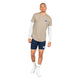 En mand står foran en hvid baggrund iført en Saysky Statement Combat Pro T-shirt, Herre, Sand, som er trukket uden på en hvid langærmet skjorte, kombineret med marineblå shorts, hvide ankelstrømper og hvide sneakers. Denne hurtigtørrende løbetrøje til herre fra Saysky er både praktisk og stilfuld, mens han står med ansigtet vendt fremad, den ene arm let bøjet, og kigger ind i kameraet.