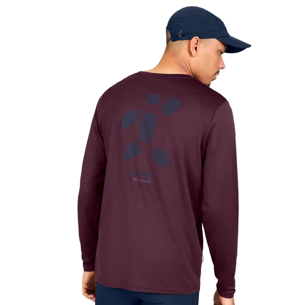 En mand iført en marineblå kasket og en langærmet »Statement Pace Long Sleeve, Herre, Burgundy« fra Saysky står med ryggen til. Bagsiden af den hurtigtørrende trøje er prydet med et enkelt, abstrakt mønster og en lille tekst. Baggrunden er hvid.