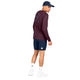 En mand står med ryggen til og har en marineblå kasket på, en Saysky Statement Pace langærmet herretrøje i farven Burgundy med et motiv på ryggen, marineblå shorts, hvide sokker med sorte stjernesymboler og hvide sneakers, alt sammen på en ensfarvet hvid baggrund. Det hurtigtørrende materiale i Saysky-trøjen sikrer komfort og ydeevne under enhver aktivitet.