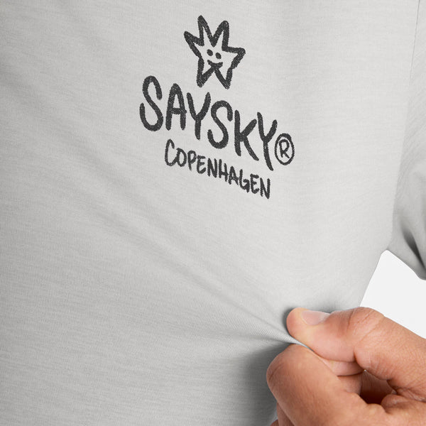 En hånd trækker i stoffet på en lysegrå Statement Pace Long Sleeve fra Saysky, der er prydet med teksten »SAYSKY Copenhagen« i sort og et stjernelogo på brystet. Denne hurtigtørrende herre-løbetrøje er designet med fokus på komfort og ydeevne.