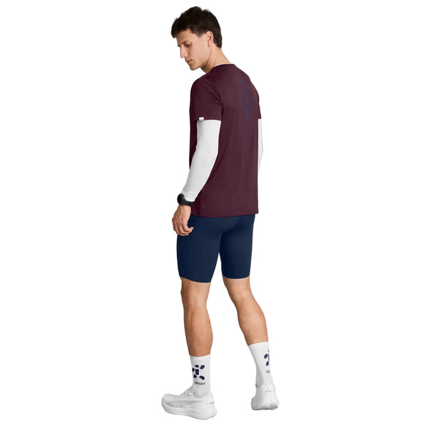 En mand står med ryggen let vendt mod kameraet og har en Saysky Statement Pace T-shirt, Herre, i farven burgundy på, over en hvid langærmet trøje, kombineret med marineblå shorts, hvide sneakers og hvide sokker med et blåt logo. Han kigger ned på sit smartwatch på venstre håndled og er klar til træning i dette afslappede herrelook.