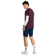 En mand står med ryggen let vendt mod kameraet og har en Saysky Statement Pace T-shirt, Herre, i farven burgundy på, over en hvid langærmet trøje, kombineret med marineblå shorts, hvide sneakers og hvide sokker med et blåt logo. Han kigger ned på sit smartwatch på venstre håndled og er klar til træning i dette afslappede herrelook.