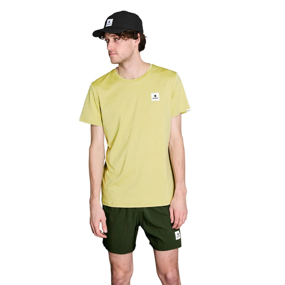 En mand iført en grøn meleret Saysky Statement Pace-T-shirt, mørkegrønne shorts og en sort kasket står foran en hvid baggrund. Alle disse beklædningsgenstande er forsynet med et lille, firkantet, hvidt logo – perfekt til træning eller fritidstøj.