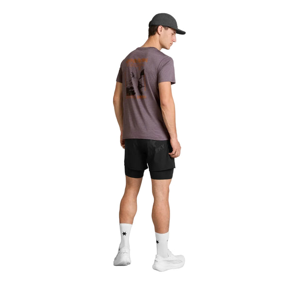 En mand står med ryggen til og har en grå kasket på, en lilla Saysky Statement Pace T-shirt til mænd med et grafisk motiv, sorte shorts, hvide sokker med sorte stjerner og hvide sneakers. Dette trendy herre-træningslook fremstår på en ensfarvet hvid baggrund.