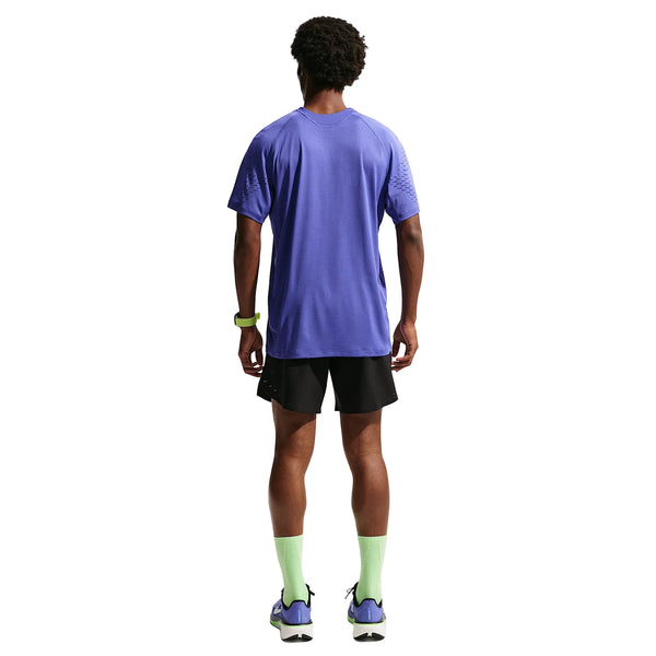 En mand ser væk fra kameraet og er iført en lilla Nike Stride Plus SS Running Top, Herre, Lapis/Deep Royal Blue/Reflective Silver, sorte shorts, lysegrønne sokker, lilla sportssko og et grønt armbånd. Baggrunden er hvid.