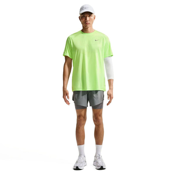 En person står med front mod en almindelig hvid baggrund iført en neongrøn Nike Stride SS Running Top (Herre, Volt Ice/Reflective Silv), grå sportsshorts, hvid kasket, sokker, sneakers og et hvidt armmanchet.