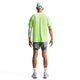En mand kigger væk fra kameraet iført en lysegrøn Nike Stride SS Running Top, Herre, Volt Ice/Reflective Silv med Dri-FIT ADV, grå shorts, hvide sokker og sneakers, en hvid kasket og et hvidt ærme på venstre arm mod en almindelig hvid baggrund.