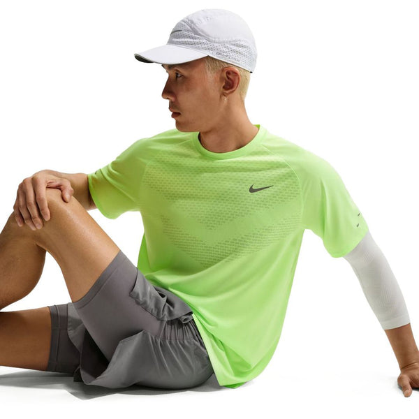 En person i en neongrøn Nike Stride SS Running Top, Herre, Volt Ice/Reflective Silv, en hvid kasket, grå shorts og et hvidt ærme sidder på jorden med det ene ben bøjet og armen på knæet mod en hvid baggrund.
