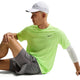 En person i en neongrøn Nike Stride SS Running Top, Herre, Volt Ice/Reflective Silv, en hvid kasket, grå shorts og et hvidt ærme sidder på jorden med det ene ben bøjet og armen på knæet mod en hvid baggrund.