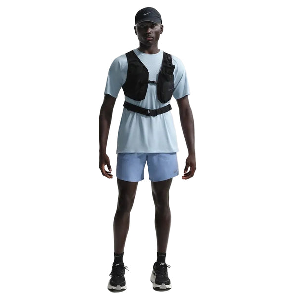 En person står med front mod en hvid baggrund iført en Nike Stride SS løbetop til mænd i Lt Armory Blue/Reflective Silver, sammen med en sort kasket, sort vest, lyseblå shorts, sorte sneakers og sorte sokker.