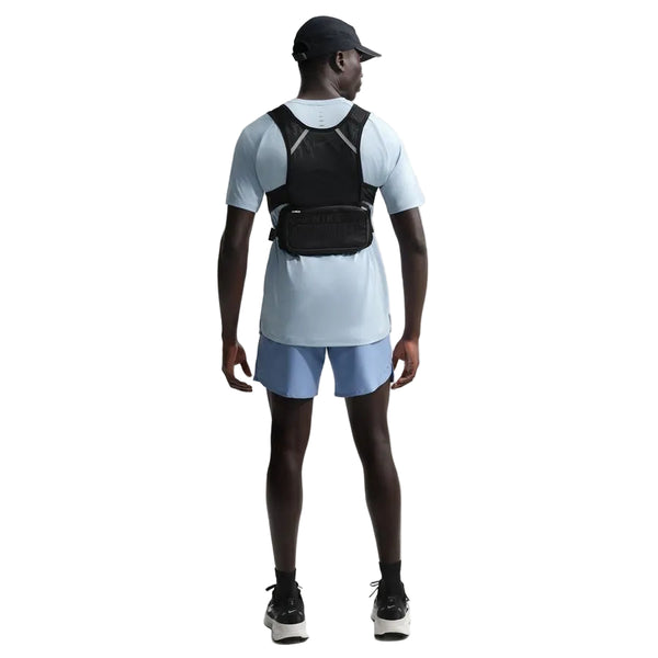 En person iført Nike Stride SS Running Top til mænd i Lt Armory Blue/Reflective Silver står med ryggen til kameraet, parret med blå shorts, sorte løbesko, en sort kasket og en sort løbevest på en hvid baggrund.