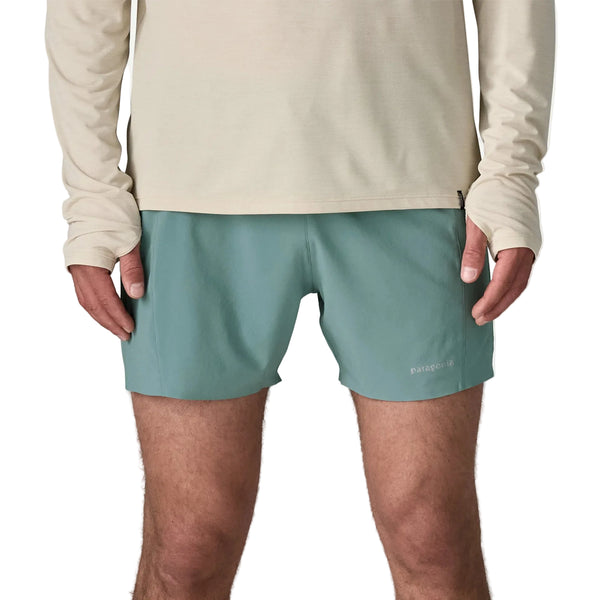 En person iført en beige langærmet skjorte og Patagonia Strider Pro Shorts 5in, Herre, Blue Sage står med front mod kameraet. Kun kroppen fra skuldre til midt på låret er synlig. Shortsene har et lille Patagonia-logo på det ene ben.