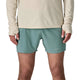 En person iført en beige langærmet skjorte og Patagonia Strider Pro Shorts 5in, Herre, Blue Sage står med front mod kameraet. Kun kroppen fra skuldre til midt på låret er synlig. Shortsene har et lille Patagonia-logo på det ene ben.