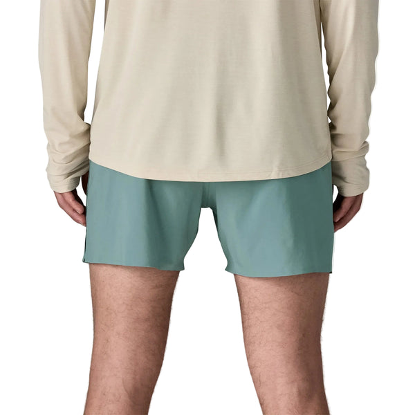En person vender ansigtet væk fra kameraet iført en langærmet beige skjorte og Patagonia Strider Pro Shorts 5in, Herre i Blue Sage. Kun den nederste del af ryggen, hofterne og benene er synlige på en hvid baggrund.