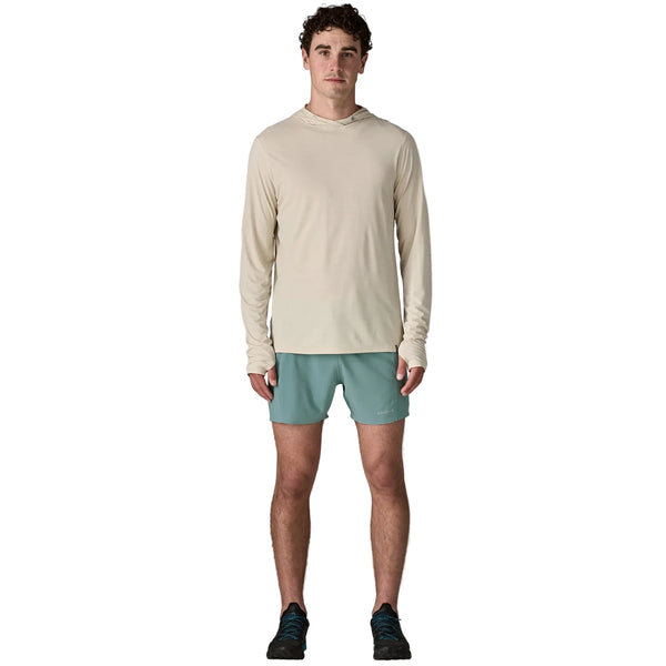 En mand står med front mod en beige langærmet hættetrøje, Patagonia Strider Pro Shorts 5in for men i Blue Sage og sorte sportssko på en almindelig hvid baggrund.