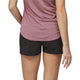 En person vises fra midt på ryggen og ned iført en lilla kortærmet skjorte og Patagonia Strider Pro Shorts 3.5", Dame, Black-hurtigtørrende træningsshorts - stående mod en almindelig hvid baggrund med venstre arm let bøjet.