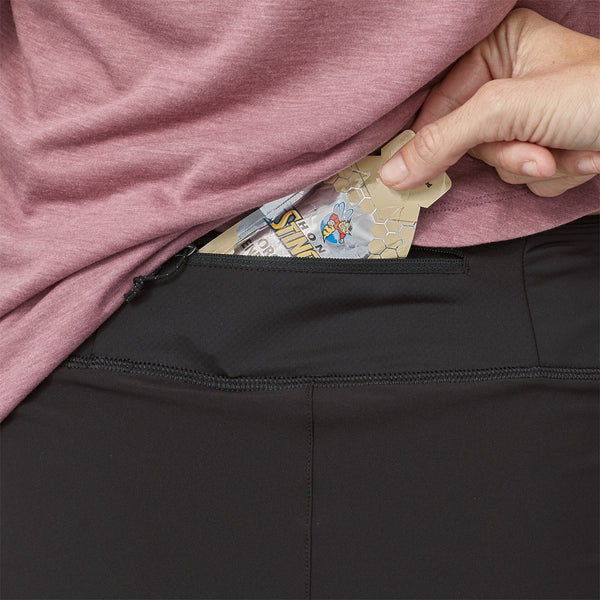 En person i lyserød skjorte og sorte bukser stikker en energigel ned i lynlåslommen på sine Patagonia Strider Pro Shorts 3,5", Dame, Black-perfekte hurtigtørrende træningsshorts til løb for damer.