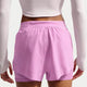 En person iført en langærmet hvid crop top og Nike Swift Df Mr 2in1 Shorts, Dame, Lt Magenta/Lt Magenta/Reflective Silv ses bagfra, stående med armene afslappet ned langs siden.
