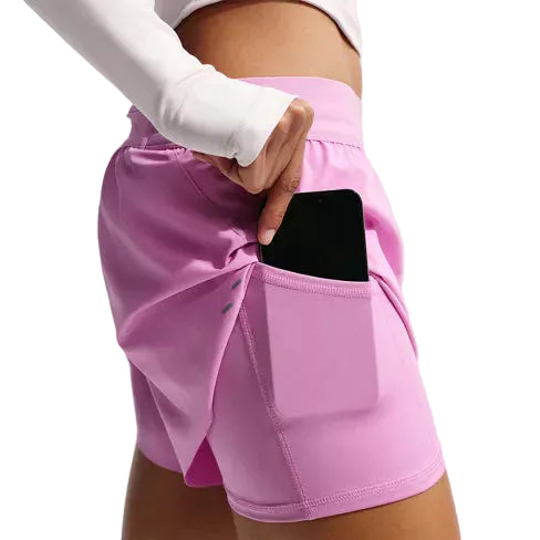 En person er iført lys magenta Nike Swift Df Mr 2in1 Shorts, Dame, med en telefon delvist gemt i sidelommen og kombinerer dem med en langærmet hvid top mod en almindelig hvid baggrund.