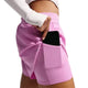 En person er iført lys magenta Nike Swift Df Mr 2in1 Shorts, Dame, med en telefon delvist gemt i sidelommen og kombinerer dem med en langærmet hvid top mod en almindelig hvid baggrund.