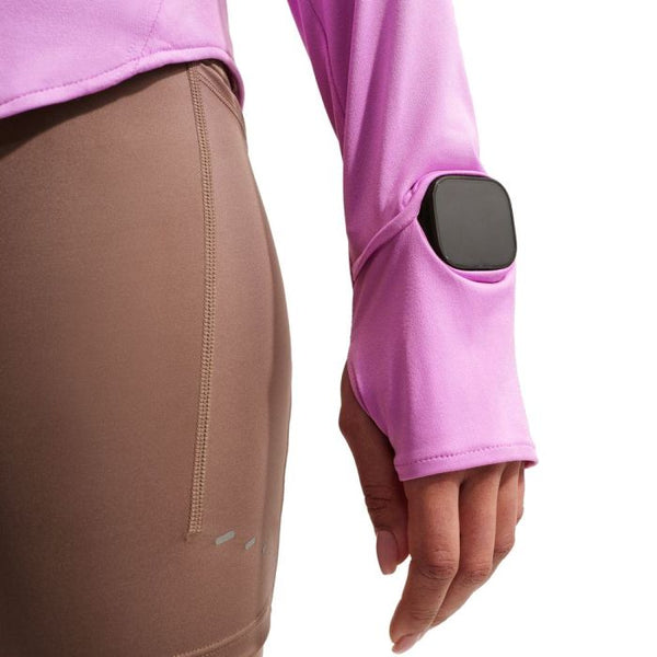 En person i brune træningsleggings og en pink Nike Swift UV Half Zip Running Top (Dame, Lt Magenta/Reflective Silv) med et smartwatch stukket ned i ærmeslidsen nær håndleddet. Kun underkroppen og armen er synlige.