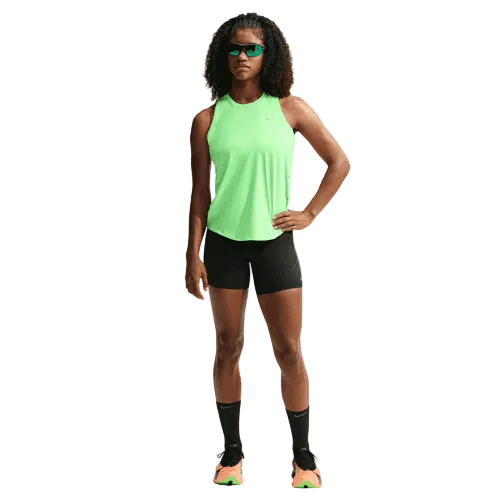 En person iført en Nike Swift Dri-FIT løbetop til kvinder i farven Illusion Green/Reflective Silver, sorte shorts og sokker, grønne sportssko og grønne solbriller står med ansigtet vendt fremad og den ene hånd på hoften.