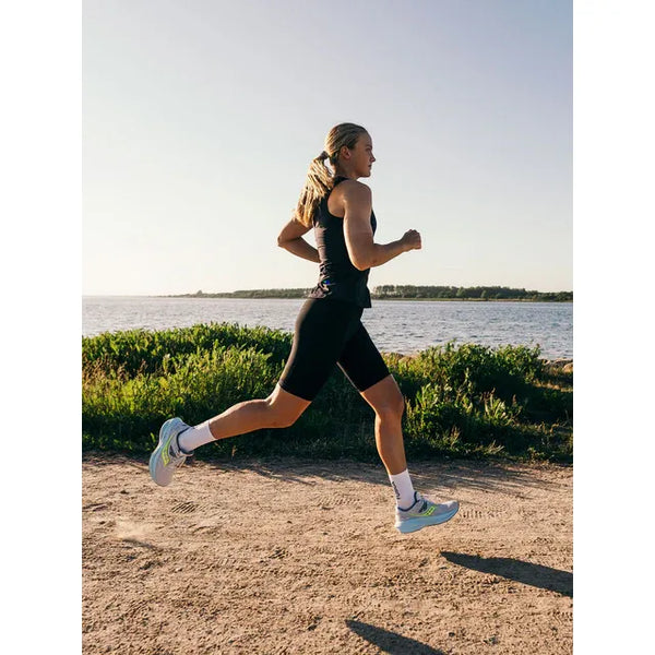 En person med langt hår i hestehale løber på en grussti ved vand iført Fusion TEMPO! Run Tights (Unisex, sort), sportstøj, hvide sokker og løbesko. Der er grønne buske og træer i baggrunden.