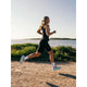 En person med langt hår i hestehale løber på en grussti ved vand iført Fusion TEMPO! Run Tights (Unisex, sort), sportstøj, hvide sokker og løbesko. Der er grønne buske og træer i baggrunden.