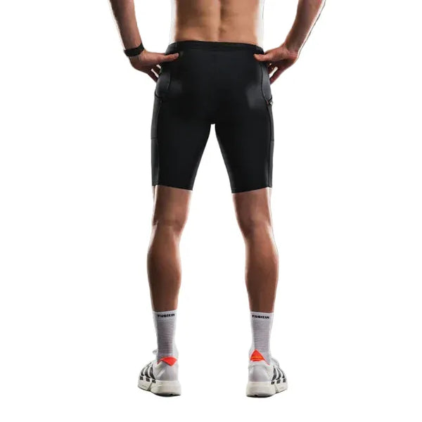 En person står med ryggen til på en hvid baggrund iført Fusion TEMPO! Run Tights (unisex, sort) sammen med et mørkt armbånd, hvide sokker med tekst og hvide sneakers med sorte striber og orange detaljer.