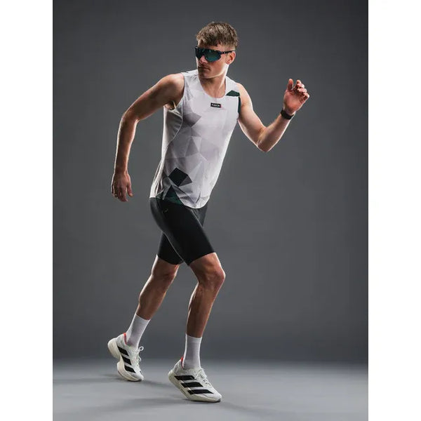En mand iført solbriller, en ærmeløs hvid top med geometrisk mønster, Fusion TEMPO! Run Tights (Unisex, Black), hvide sokker og hvide sneakers står i en løbepositur mod en almindelig grå baggrund.