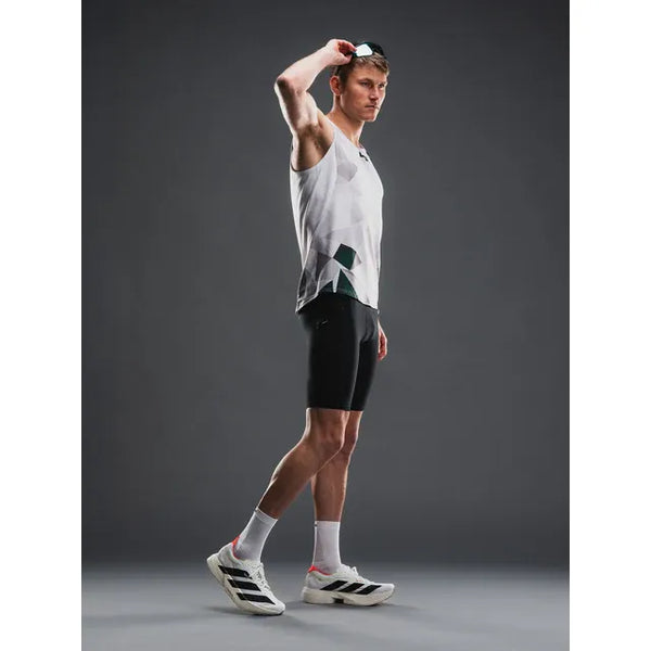 En mand i sportstøj står på en grå baggrund iført en hvid og grå tanktop, Fusion TEMPO! Run Tights (Unisex, Black), hvide sokker og hvide løbesko, mens han løfter solbrillerne af hovedet med den ene hånd.