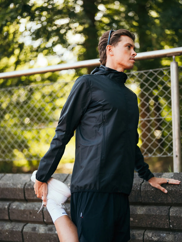 En person iført Fusion Tempo! Two Run-jakken (herre, sort) og shorts strækker benet ved at holde foden bag sig udendørs tæt på en stenmur og et metalhegn, med træer i baggrunden.