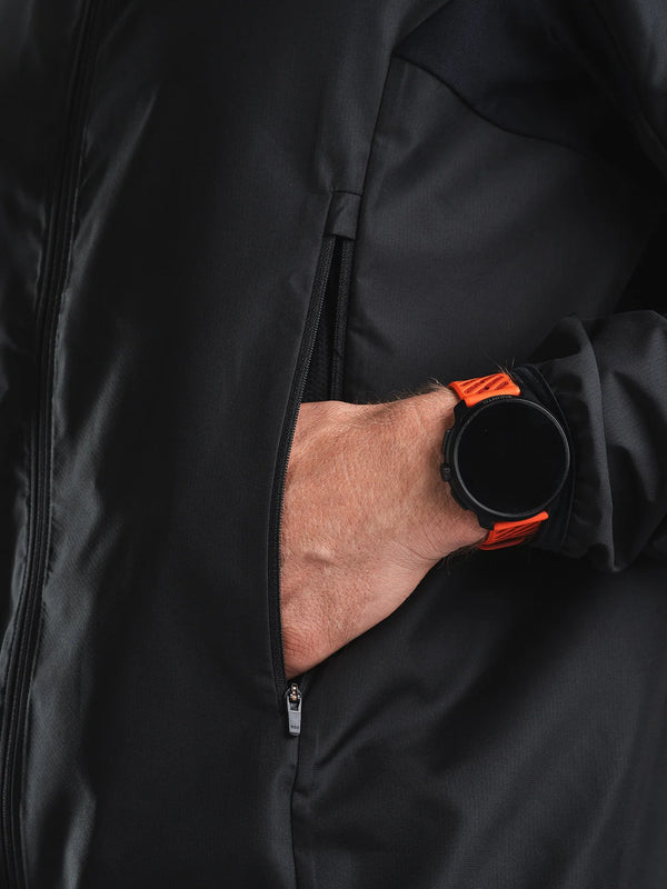 En person, der har Fusion Tempo! Two Run-jakken på (herre, sort) og har hånden i lommen, bærer desuden et rundt, sort smartwatch med et orange armbånd.