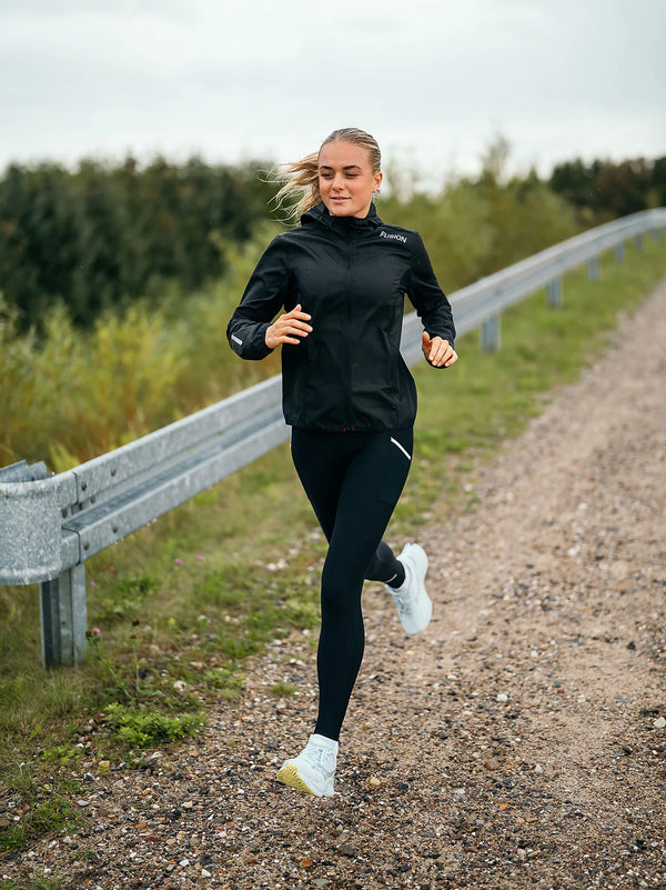 En kvinde iført en sort Fusion Tempo! Two Run-jakke og leggings løber ad en grusvej.