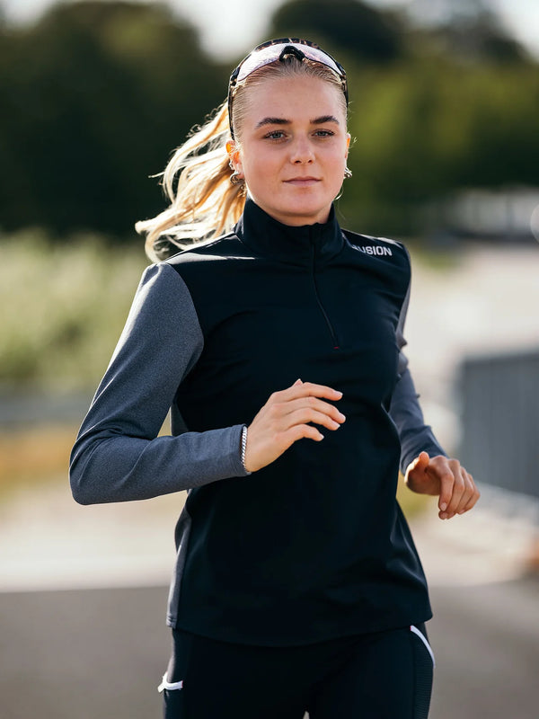 En person med langt blondt hår, iført en Fusion Tempo! Shield Zip hybrid-løbetrøje til kvinder i mørkegrå og solbriller på hovedet, løber udendørs i dagslys foran en sløret baggrund af grønne omgivelser.