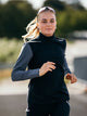 En person med langt blondt hår, iført en Fusion Tempo! Shield Zip hybrid-løbetrøje til kvinder i mørkegrå og solbriller på hovedet, løber udendørs i dagslys foran en sløret baggrund af grønne omgivelser.