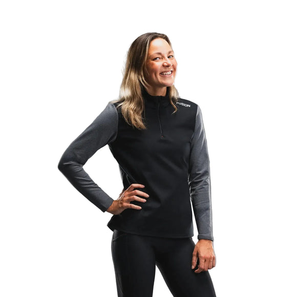En kvinde står foran en hvid baggrund og smiler med den ene hånd på hoften. Hun har Fusion Tempo! Shield Zip, Dame, mørkegrå på – en aktiv, komfortabel og funktionel langærmet bluse med ordet »FUSION« tæt på venstre skulder – kombineret med sorte bukser.
