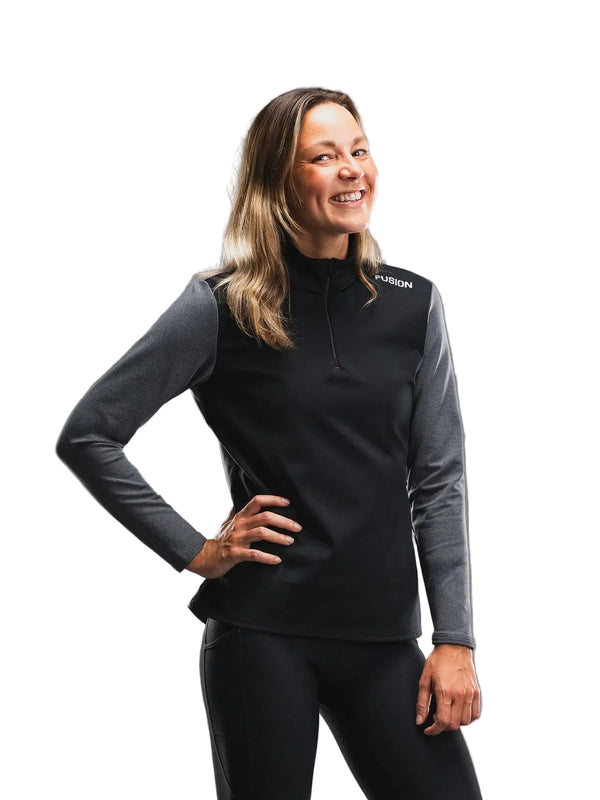 En kvinde står foran en hvid baggrund og smiler med den ene hånd på hoften. Hun har Fusion Tempo! Shield Zip, Dame, mørkegrå på – en aktiv, komfortabel og funktionel langærmet bluse med ordet »FUSION« tæt på venstre skulder – kombineret med sorte bukser.