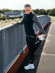 En kvinde iført Fusions »Tempo! Shield Zip«-jakke i mørkegrå står på en løbebane. Hun er klædt i sort træningstøj og hvide sneakers og holder fast i et gelænder med den ene hånd, mens hun strækker benet bagud. Den hybrid-løbetrøje sikrer, at hun er forberedt på al slags vejr, og hun har solbrillerne siddende på hovedet. I baggrunden ses træer og bygninger.